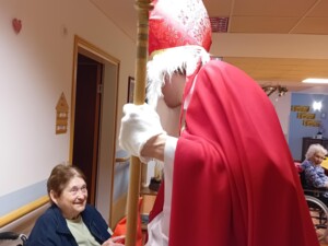 Eine ältere Person im Rollstuhl nimmt freudig ein Geschenk vom Nikolaus entgegen. Sie sitzt an einem gedeckten Tisch im Gemeinschaftsbereich des Pflegeheims.