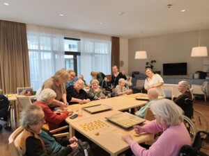Eine größere Gruppe von Bewohner:innen sitzt und steht um einen Tisch und formt gemeinsam Kekse. Auf Backblechen liegen bereits vorbereitete Teigstücke. Betreuungspersonen unterstützen die Gruppe. Der helle Raum und die gemeinschaftliche Aktivität vermitteln eine lebendige und herzliche Stimmung.