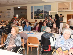 Mehrere Senior:innen sitzen an weiß gedeckten Tischen in der Eingangshalle. Einige tragen bunte Hüte oder Faschingsaccessoires, manche nutzen Rollstühle oder Rollatoren. Gläser mit Getränken stehen auf den Tischen. Im Hintergrund klatschen Mitarbeitende und Besucher:innen, während die Stimmung lebhaft und fröhlich wirkt.