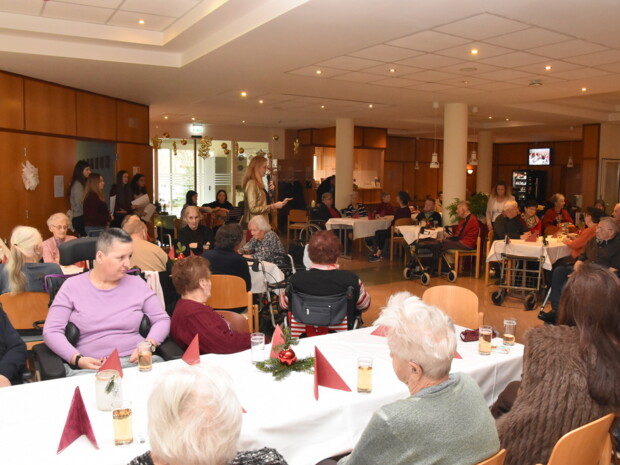 Zahlreiche Senior:innen und Gäste sitzen an festlich gedeckten Tischen in der Eingangshalle des Heims St. Franziskus. Eine Frau hält ein Mikrofon und spricht in die Runde, während im Hintergrund junge Musiker:innen stehen. Haus- und Pflegedienstleiterin Julia Preissecker spricht Begrüßungsworte.