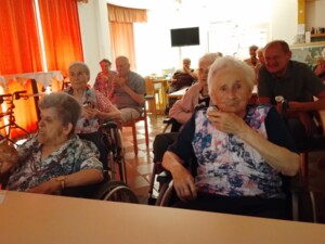 Mehrere ältere Bewohner:innen sitzen in einem Aufenthaltsraum, teils in Rollstühlen, und essen Eis. Rote Vorhänge schützen vor der Sonne, im Hintergrund ist eine kleine Bar zu sehen.