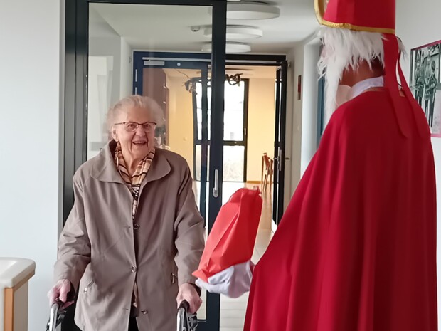 Eine ältere Person mit Rollator begegnet dem Nikolaus auf dem Flur eines Pflegewohnhauses. Beide lachen, und der Nikolaus hält ein rotes Geschenk in der Hand.