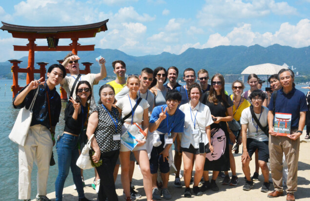 Eine diverse Gruppe von etwa zwanzig Personen posiert bei sonnigem Wetter für ein Gruppenfoto vor dem roten Torii des Itsukushima-Schreins auf der Insel Miyajima in Japan. Im Hintergrund sind das Meer, grüne Berge und ein blauer Himmel mit weißen Wolken zu sehen. Einige Personen tragen Namensschilder, was auf eine organisierte Reise oder ein Austauschprogramm hinweist.