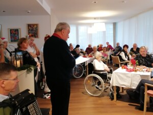 Ein festlich dekorierter Raum ist mit Bewohner:innen gefüllt, die an Tischen sitzen. Eine Person steht vorne und hält eine Ansprache, während daneben eine weitere Person ein Akkordeon spielt. Im Hintergrund stehen Mitarbeitende und hören zu. Die Atmosphäre wirkt feierlich und gemeinschaftlich.