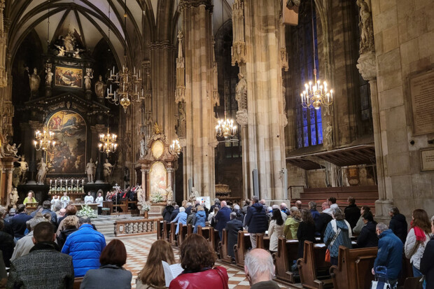 Zahlreiche Menschen nehmen an einem Gottesdienst im Stephansdom in Wien teil. Sie sitzen und stehen in den Kirchenbänken, während vorne Geistliche am Altar die Messe zelebrieren. Der gotische Innenraum ist reich mit Skulpturen, Altären und Gemälden verziert. Kronleuchter erhellen den Raum, und im Hintergrund ist ein Chor zu sehen.