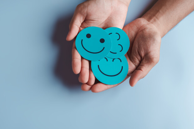 Symbolbild. Zwei geöffnete Hände halten drei türkisfarbene runde Karten mit verschiedenen Smileys, die Freude und positive Emotionen ausdrücken. Der Hintergrund ist einfarbig hellblau.