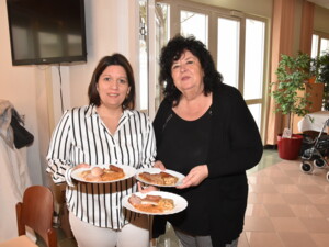 Zwei Senior:innenbetreuerinnen stehen in einem festlich dekorierten Raum und halten mehrere Teller mit Speisen wie Geselchtem, Würsteln, Sauerkraut und Knödeln.