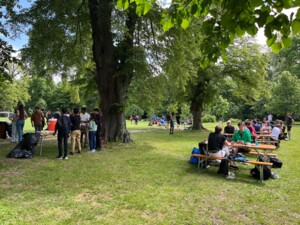 Menschen sitzen an Bierzeltgarnituren unter Bäumen und essen oder unterhalten sich. Links im Bild stehen andere Personen an einem Getränketisch. Im Hintergrund sind Fußballspiele im Gange.