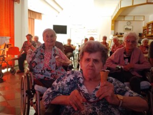 Eine Gruppe älterer Bewohner:innen sitzt im Aufenthaltsraum, mehrere Personen halten Eis in der Hand. Einige nutzen Rollstühle oder Gehhilfen, links fallen rote Vorhänge ins Bild.