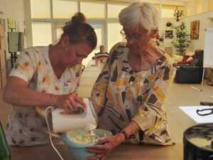 Eine Mitarbeiter:in unterstützt eine Bewohner:in beim Rühren von Teig mit einem Handmixer in einer blauen Schüssel. Beide stehen an einem Tisch im Gemeinschaftsraum, im Hintergrund sind weitere Bewohner:innen und Sitzbereiche zu sehen.
