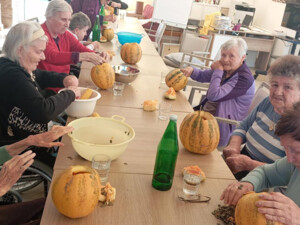 Eine Gruppe älterer Menschen sitzt an einem langen Tisch. Jede Person bearbeitet einen Kürbis, um Kerne zu entfernen oder ihn vorzubereiten. Der Tisch ist mit Schüsseln und Gläsern gedeckt.