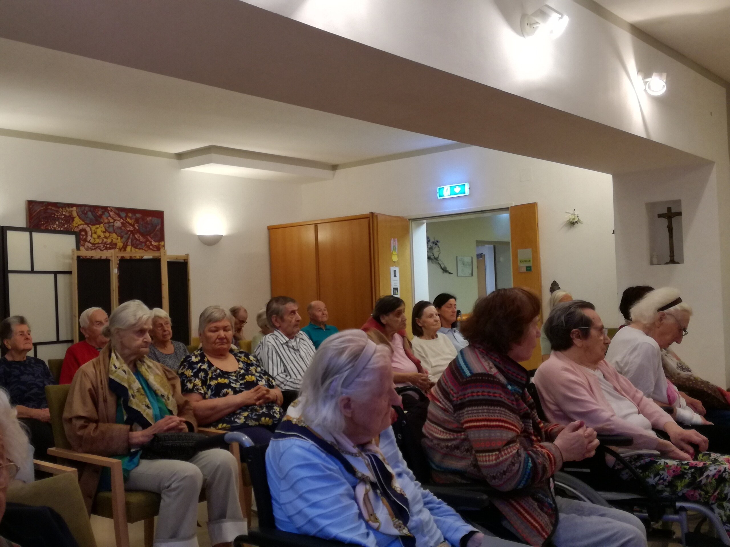 Mehrere Senior:innen sitzen aufmerksam in Reihen in einem hell erleuchteten Raum mit religiöser Symbolik an den Wänden. Sie nehmen an einer heiligen Messe in der Hauskapelle der Einrichtung Aigen teil. Die Teilnehmenden wirken ruhig und andächtig, einige nutzen Rollstühle. Die Veranstaltung fand am 18. September statt.
