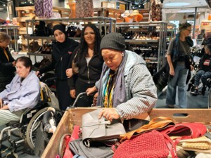 Mehrere Bewohner:innen und Begleiter:innen befinden sich in einer Halle mit Regalen voller Secondhand-Waren wie Schuhe und Taschen. Im Vordergrund durchsucht eine Person eine Kiste mit Handtaschen, während andere aufmerksam zusehen. Einige Personen sitzen im Rollstuhl. Die Szene zeigt gemeinsames Entdecken und lebendiges Interesse an den angebotenen Gegenständen.