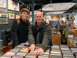 Zwei Personen stehen in einem Secondhand-Geschäft vor einem Tisch mit zahlreichen CDs. Eine Person blättert durch die Sammlung, während die andere danebensteht. Im Hintergrund sind weitere Regale mit unterschiedlichen gebrauchten Artikeln zu sehen, darunter Sportgeräte und Bilder.