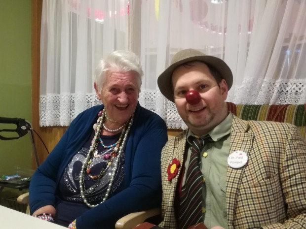 Bewohnerin und Clown sitzend nebeneinander fröhlich in die Kamera schauend.