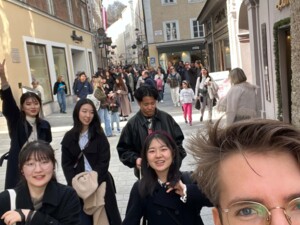 Sechs Personen in Winterkleidung gehen in einer engen Straße mit alten Häusern auf die Kamera zu. Das Bild scheint spontan als Selbstportrait geschossen worden zu sein: Nicht alle Personen blicken in die Kamera, manche sind nur teilweise abgeschnitten und das Bild ist nicht ganz gerade. Der Baustil der weiß und gelb verputzten Häuser im Hintergrund und die daran angebrachten, in die Gasse ragenden Metallschilder- und -laternen lassen auf einen Ort in Europa schließen. Im Hintergrund sind viele Menschen zu sehen. Das Wetter ist sonnig.