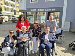 Mehrere ältere Menschen in Rollstühlen sowie stehende Begleitpersonen posieren gemeinsam vor einem Gebäude mit der Aufschrift "Foto Pöschlmayer". Die Gruppe lächelt und wirkt zufrieden, während sie den Markttag gemeinsam verbringt.