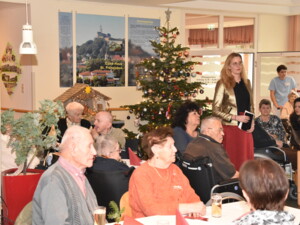 Ein geschmückter Weihnachtsbaum und eine Krippe stehen im Hintergrund. Bewohner:innen in Rollstühlen und auf Stühlen hören der Rede einer Frau im goldenen Blazer zu.
