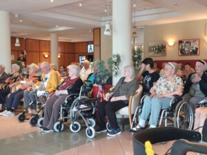 Viele Bewohner:innen sitzen in einem großen Gemeinschaftsraum, teils in Rollstühlen oder mit Rollatoren. Sie blicken nach vorne und hören der musikalischen Darbietung zu.