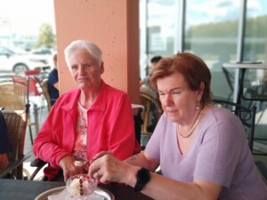 Zwei Personen sitzen an einem Tisch im Außenbereich des Café Milan. Vor ihnen steht eine Eisschale mit Eis. Eine Person trägt eine rote Jacke, die andere ein hellviolettes Shirt und reicht gerade einen Löffel weiter.