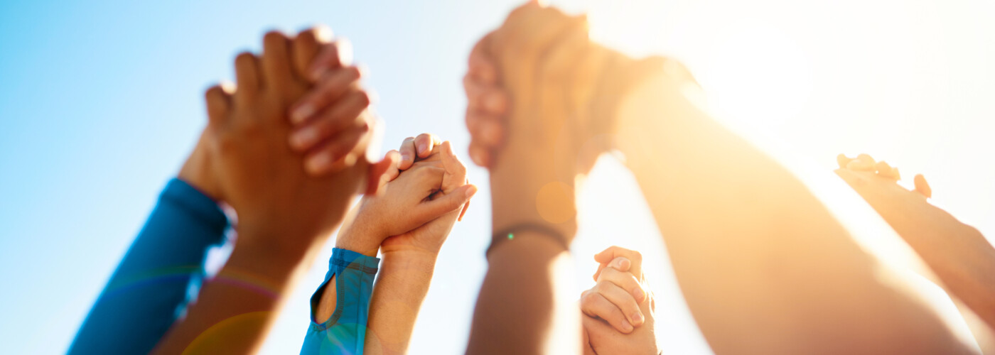 Mehrere Personen halten sich an den Händen und strecken diese vereint in den Himmel. Die Szene ist bei Tageslicht aufgenommen, mit Sonnenlicht, das von hinten durch die Arme scheint. Die Geste steht für Solidarität, Teamgeist und gemeinsames Engagement.