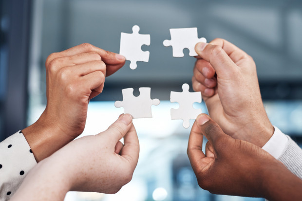 Symbolbild Vier Hände von Personen mit unterschiedlicher Hautfarbe halten jeweils ein weißes Puzzlestück hoch, die sich in der Mitte zu einem Ganzen ergänzen. Die Szene symbolisiert Diversität, Kooperation und gemeinsames Problemlösen.