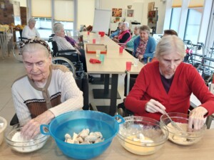 Zwei ältere Personen panieren Pilzstücke in verschiedenen Schüsseln mit Mehl, Ei und Bröseln. Weitere Bewohner:innen sitzen im Hintergrund an einem langen Tisch.