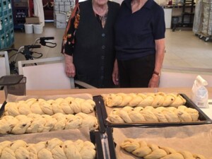 Zwei Senior:innen stehen nebeneinander hinter einem Tisch mit mehreren fertig geflochtenen Striezeln auf Backblechen. Sie lächeln in die Kamera.