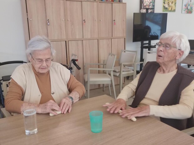 Zwei ältere Bewohnerinnen sitzen nebeneinander an einem Tisch und rollen jeweils ein Stück Teig mit den Händen zu länglichen Strängen, um daraus Fastenbeugel zu formen. Auf dem Tisch stehen ein Glas Wasser und ein grüner Kunststoffbecher. Im Hintergrund sind Holzschränke, Bilder mit Blumenmotiven und ein Fernseher im Aufenthaltsraum zu sehen.