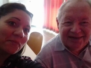 Zwei Personen sitzen nebeneinander und lächeln in die Kamera. Im Hintergrund sind rote Vorhänge und ein gedeckter Tisch zu sehen.
