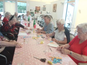 Eine Gruppe älterer Bewohner:innen sitzt gemeinsam mit Betreuungspersonen an einem langen Tisch und bastelt mit Papier, Schere und Kleber farbenfrohe Dekorationen in Form von Eiswaffeln. Die Atmosphäre wirkt konzentriert und zugleich herzlich und fröhlich.