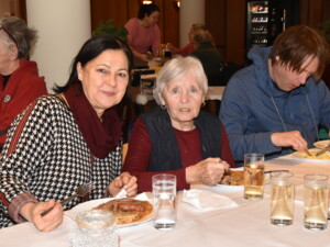 Drei Personen, darunter eine ältere Frau und zwei Begleitpersonen, sitzen an einem Tisch und genießen das Mittagessen. Im Hintergrund weitere Gäste des Festes.
