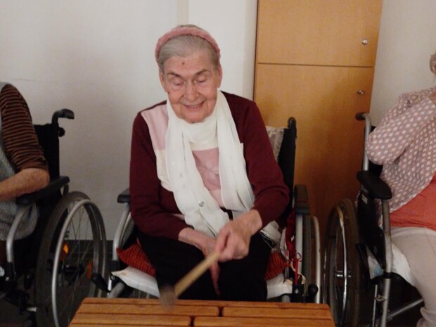 Eine ältere Person im Rollstuhl spielt mit einem Schlägel auf einem Klanginstrument aus Holz. Die Person lächelt und wirkt konzentriert. Rechts daneben sitzt eine weitere Person, die aus einem Becher trinkt. Die Szene findet in einem hellen Innenraum statt.
