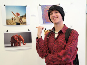 Ein jugendlicher Teilnehmer mit kariertem Hemd und Mütze steht vor einer weißen Wand mit KI-generierten Bildern. Er zeigt lächelnd auf sein eigenes Werk - ein digital erstellter Elefant, dessen Körper aus glänzenden Erdbeeren besteht.