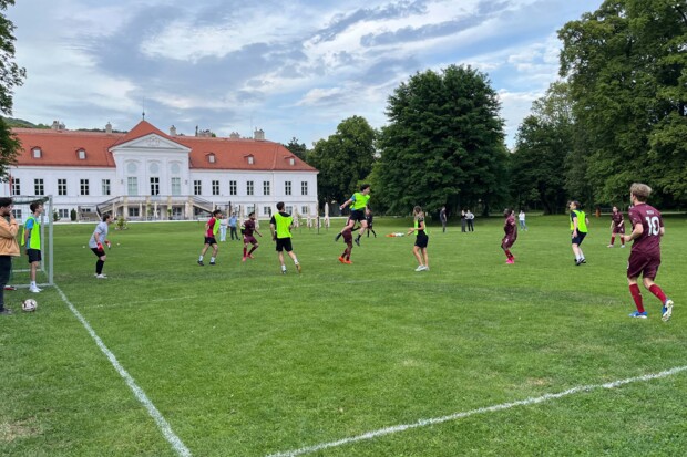 Fußballszene direkt vor dem Tor mit mehreren Spieler:innen in grünen und weinroten Trikots. Eine Person springt zum Kopfball. Im Hintergrund ein historisches Gebäude mit rotem Dach und mehrere Zuschauer:innen auf der Wiese.