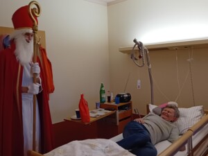 Ein älterer Mensch liegt lächelnd in einem Pflegebett, während eine als Nikolaus verkleidete Person mit rotem Mantel und Bischofsstab danebensteht. Auf dem Tisch neben dem Bett liegt ein rotes Geschenk.