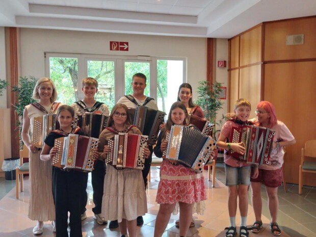 Eine Gruppe von Schüler:innen und Musikschullehrerin Elisabeth Unger steht mit Ziehharmonikas in einem hellen Aufenthaltsraum des ÖJAB-Hauses St. Franziskus. Die Musiker:innen lächeln in die Kamera.