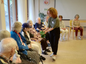 Lehrerin zeigt Senior gerade Instrument. Daneben SeniorInnen fröhlich sitzendim Stuhlkreis im Festsaal des Hauses.