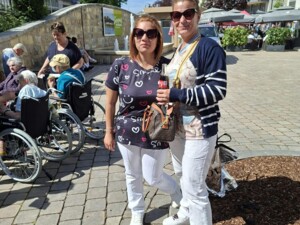 Zwei Frauen stehen auf einem gepflasterten Platz und schauen in die Kamera. Eine hält eine Getränkeflasche. Im Hintergrund sind weitere Teilnehmende des Ausflugs sowie Marktstände und Gebäude sichtbar.